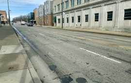 Photo of street next to buildings. The street shows patching and wear and tear.