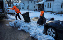 City crews in orange reflective jackets carrying trash bags through deep snow