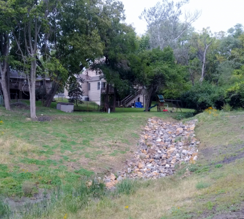 Photo of rip rap along new drainage ditch surrounded by green grass, trees and behind homes